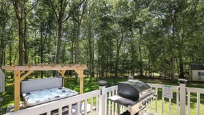 An outdoor deck with a hot tub and a grill, surrounded by a wooden fence and a wooded area. - Premium Restored Cabin | Pocono Mountains (Barrett Township)