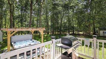 An outdoor deck with a hot tub and a grill, surrounded by a wooden fence and a wooded area.