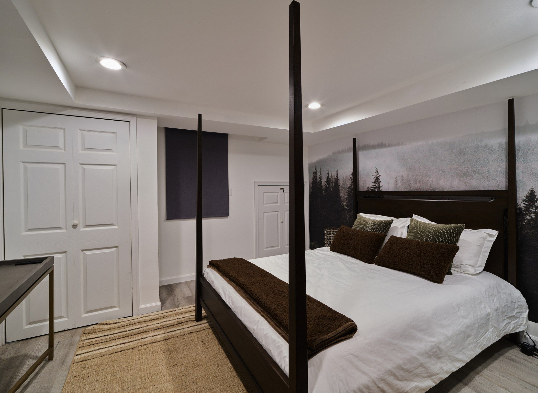 A bedroom with a four-poster bed, a large window with a forest view, and a door with a dark panel.