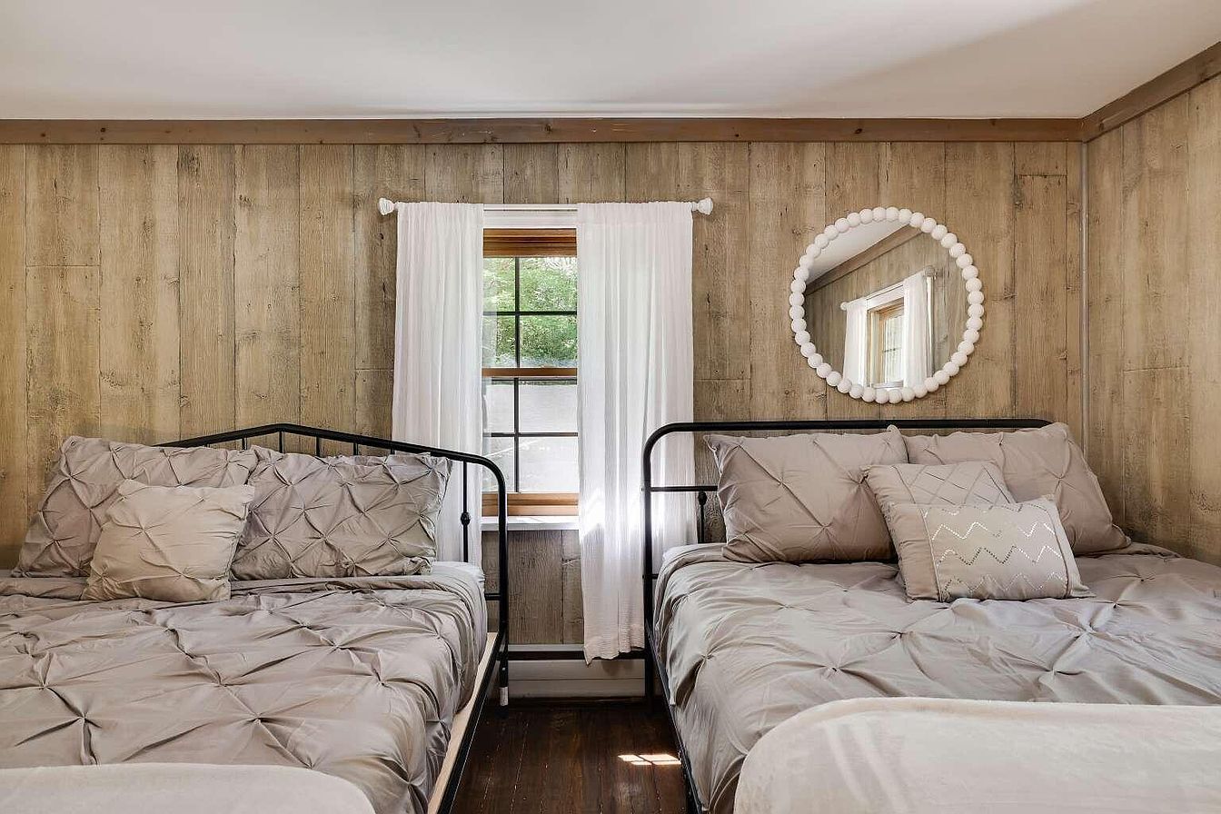 Un dormitorio con dos camas, paredes de madera, una ventana con cortinas y un espejo en la pared.