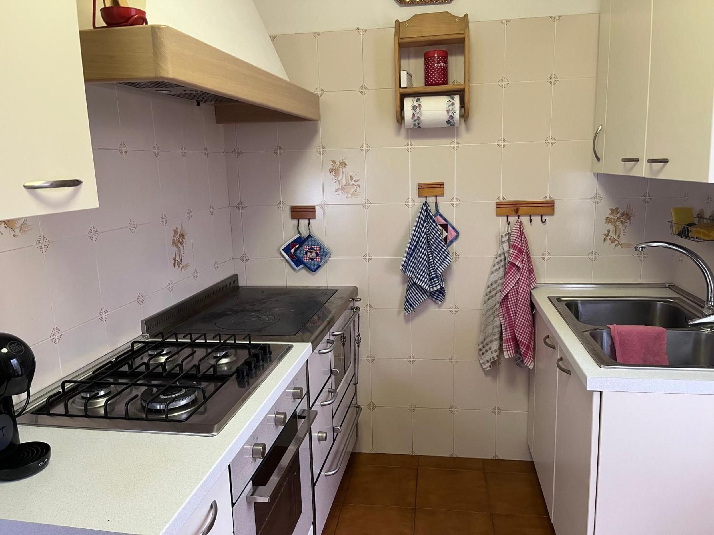 A kitchen with a gas stove, oven, and a sink with a faucet. There are hanging towels and a coffee cup holder on the wall.