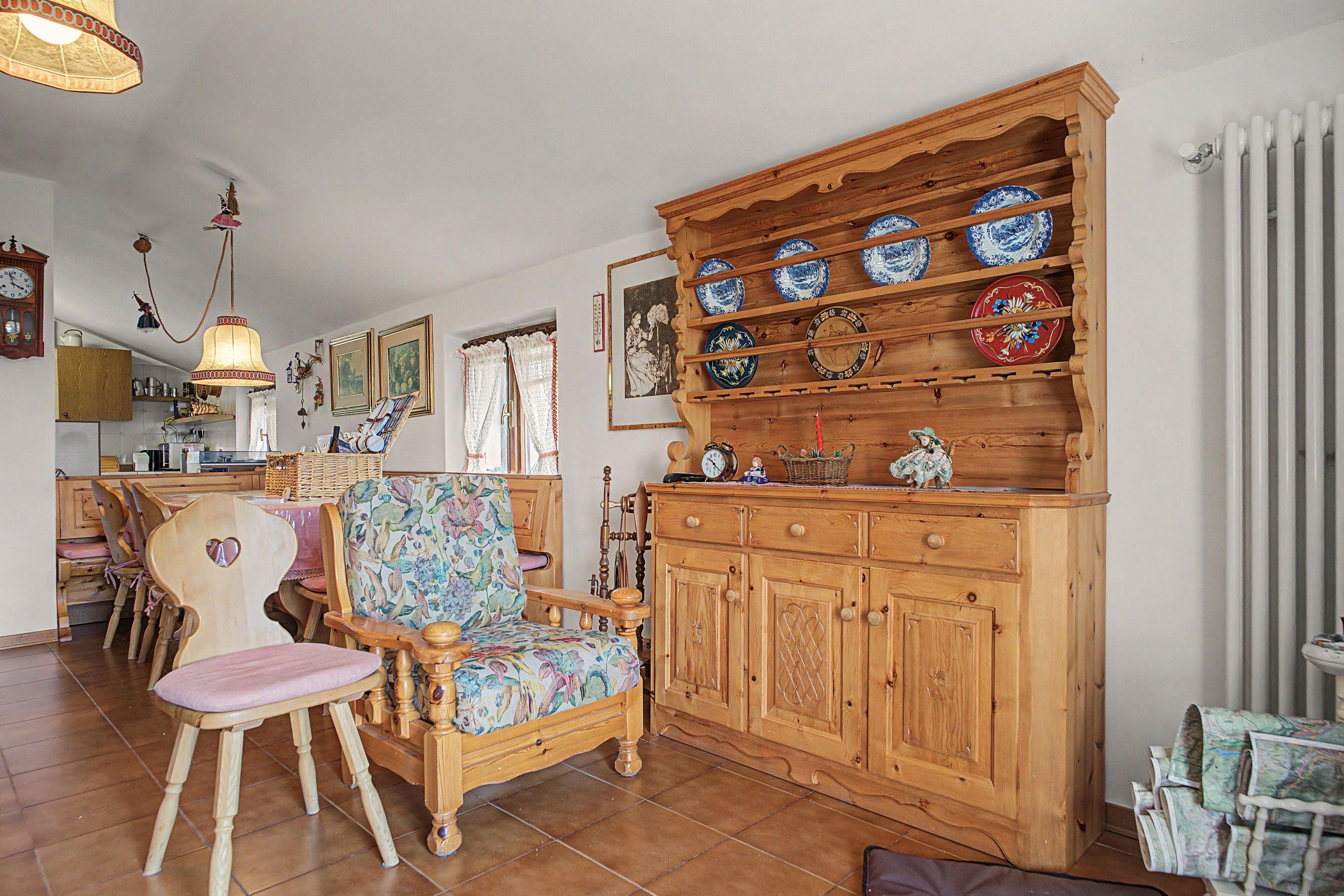 Un intérieur traditionnel avec un meuble en bois où sont exposées des assiettes en céramique, un fauteuil recouvert d’un tissu à motifs floraux et une horloge ancienne.