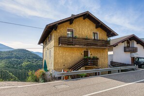 A two-story chalet with a wooden balcony, a staircase leading to the entrance, and a mountain backdrop.