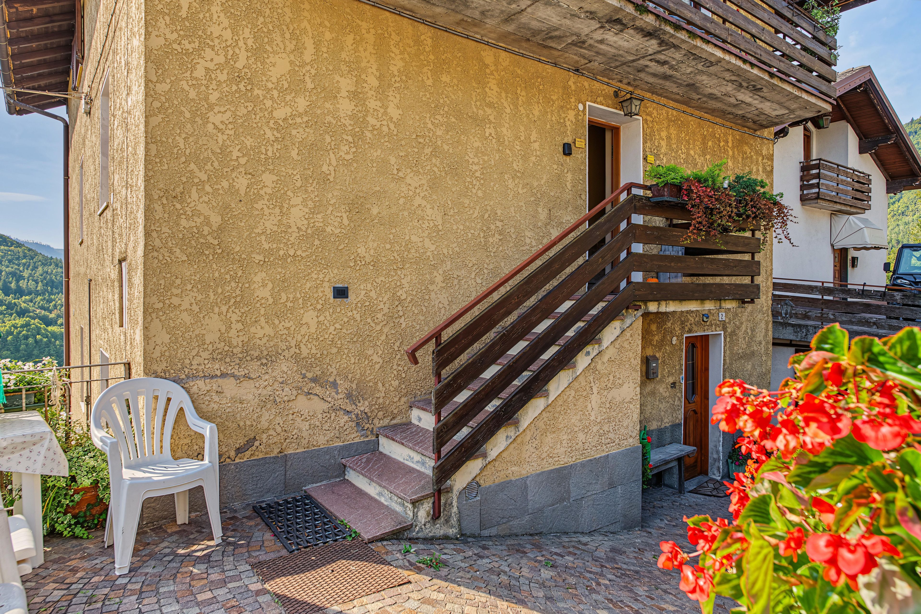 Un bâtiment avec un mur jaune à la texture particulière, un escalier en bois, une chaise en plastique blanche et une fleur rouge au premier plan.