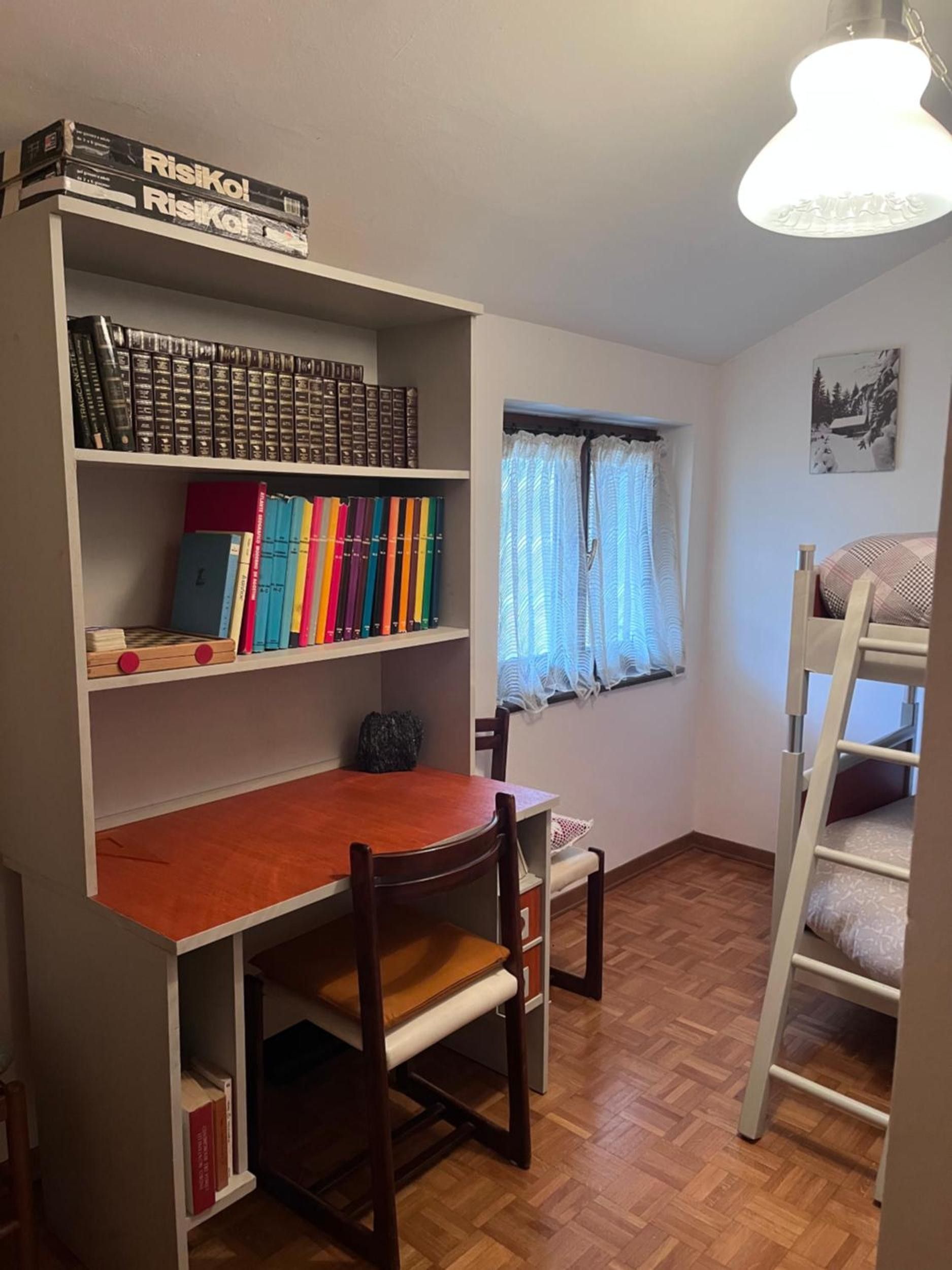 A room with a wooden desk, a bookshelf, a bunk bed, and a window with curtains.
