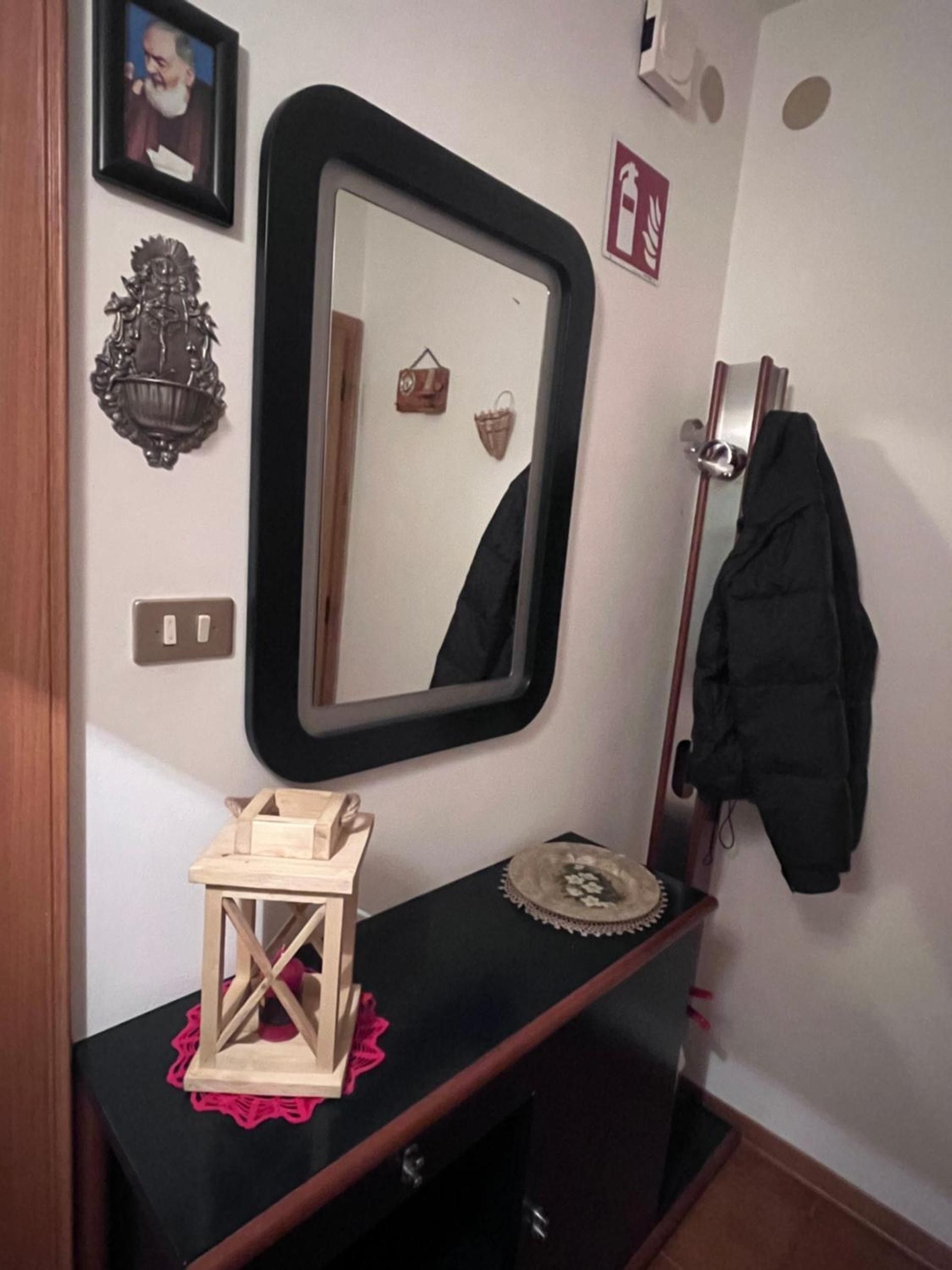 A room with a mirror, a small wooden table with a decorative item, a coat rack with a jacket, and a wall-mounted fire alarm.