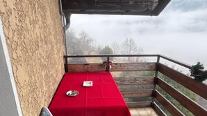 Ein Balkon mit einem roten Tisch, einem blumengedeckten Stuhl und Blick auf Bäume und Himmel.