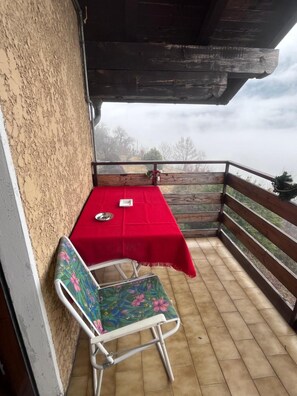 Ein Balkon mit einem roten Tisch, einem blumengedeckten Stuhl und Blick auf Bäume und Himmel.