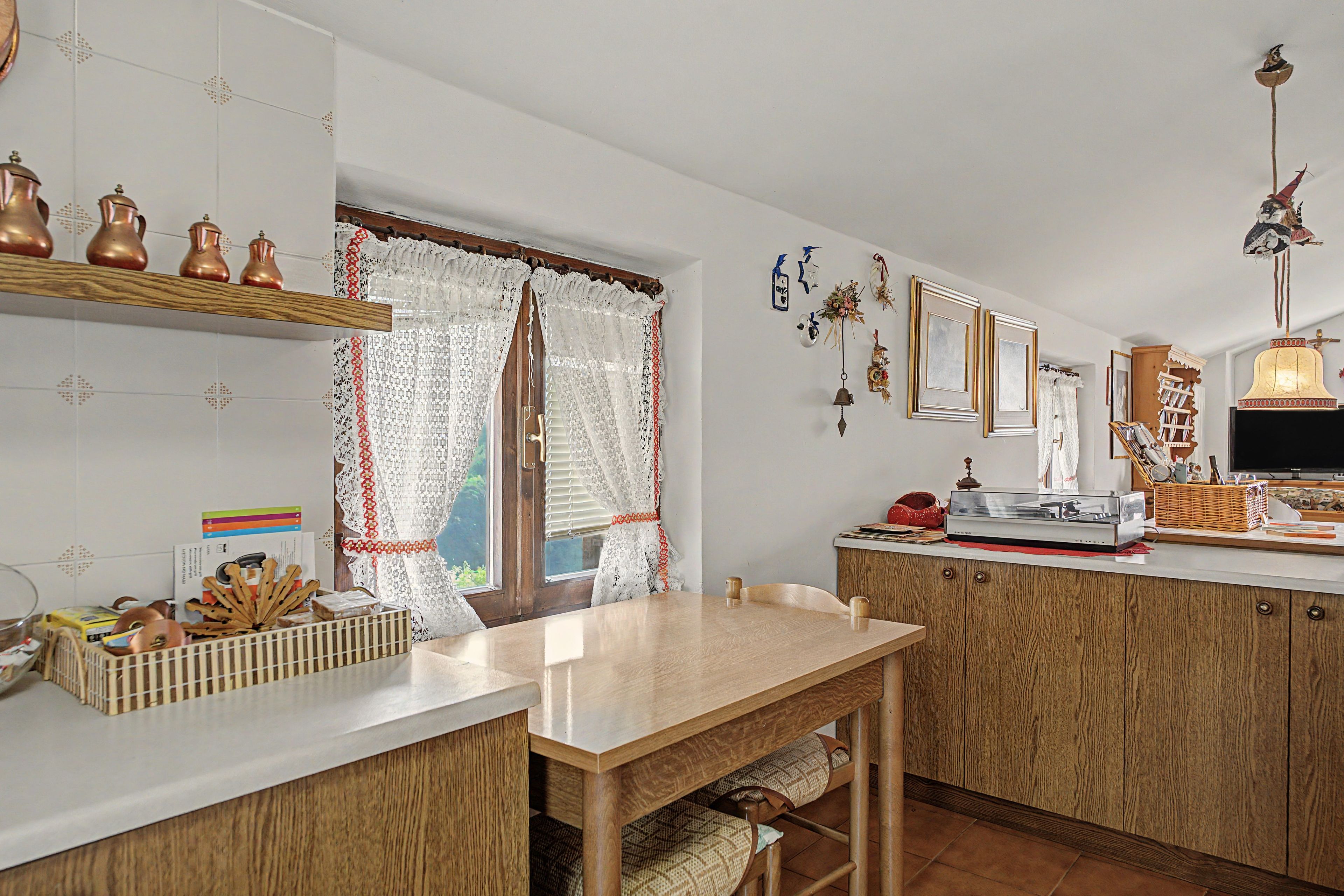 Une cuisine avec des meubles de rangement en bois, une table en bois et une fenêtre ornée de rideaux en dentelle.