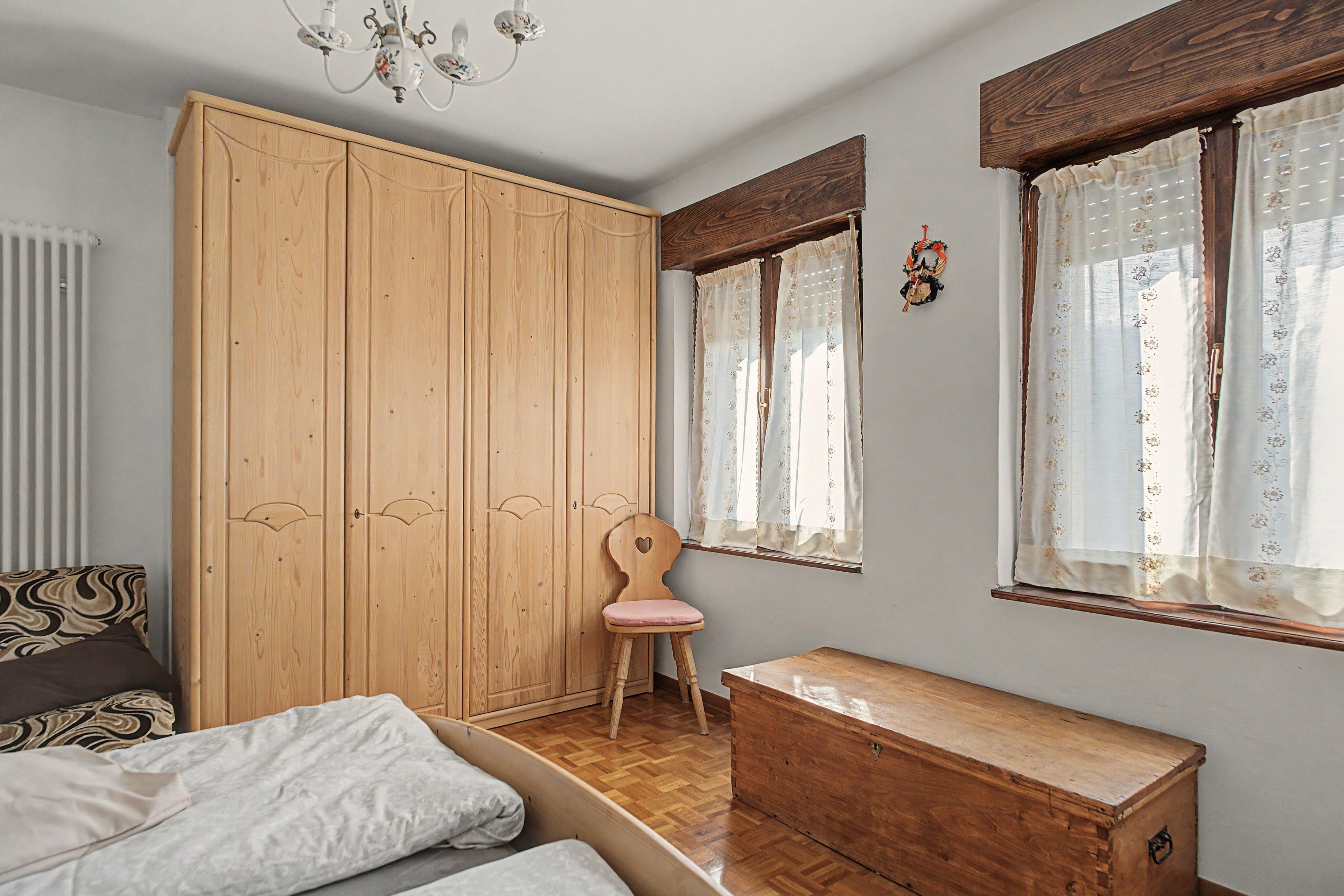 Une chambre avec une armoire en bois, une commode en bois, un lit avec une literie à motifs, une chaise et une fenêtre ornée de rideaux à fleurs.