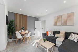 A modern living room with a dark grey sofa, white and orange cushions, a wooden coffee table, and a dining area with a white table and chairs. - Stylish 2-Bed House Near Leeds Centre | Parking (Leeds)