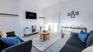 Een moderne woonkamer met een flatscreen televisie, een ronde salontafel en een eethoek met een witte tafel en stoelen.