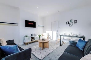 Ein modernes Wohnzimmer mit Flachbildfernseher, rundem Couchtisch und Essbereich mit weißem Tisch und Stühlen.