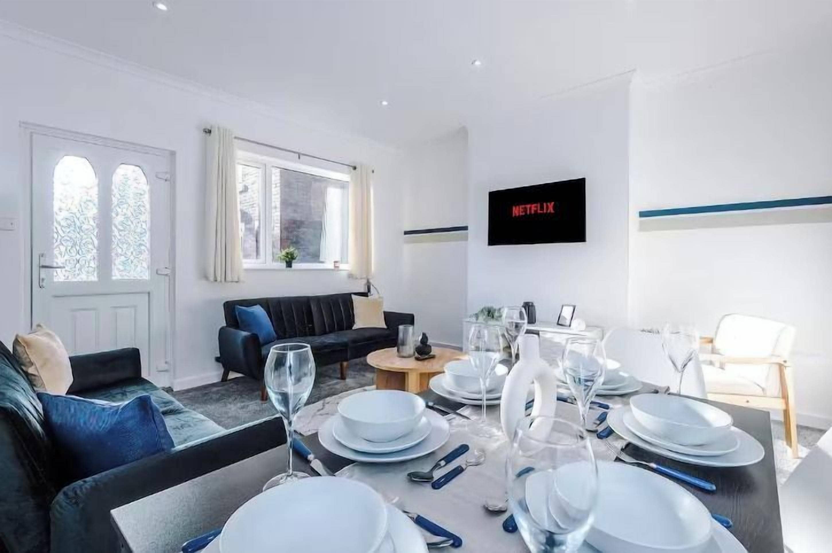 Una sala moderna con comedor listo para una comida, un sofá, un televisor montado en la pared y una ventana con cortinas.