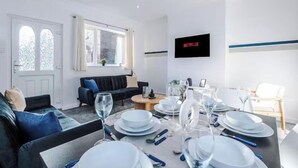 Een moderne woonkamer met een eethoek, gedekt voor een maaltijd, een bank, een aan de muur bevestigde televisie en een raam met gordijnen.