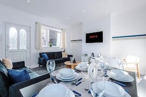 Ein modernes Wohnzimmer mit Essbereich, gedecktem Tisch, einer Couch, einem an der Wand befestigten Fernseher und einem Fenster mit Vorhängen.