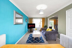 A living room with a television displaying 'NETFLIX,' a sofa with yellow cushions, and a coffee table. - Elegant 4 Bed House - Parking and Garden -Sleeps 8 (Maidstone)