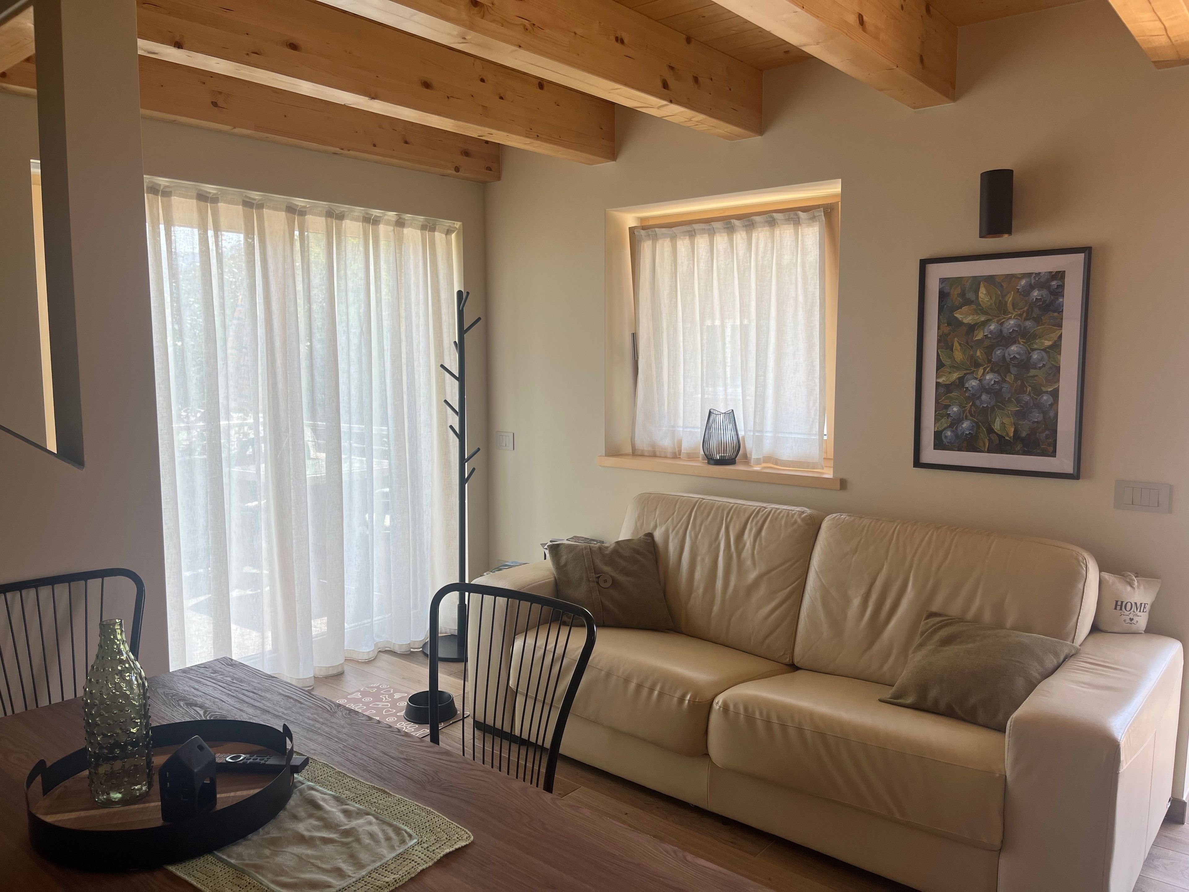 Ein Wohnzimmer mit einem beigen Sofa, einem Esstisch mit Stühlen, einer Holzbalkendecke und einem Fenster mit Vorhang.