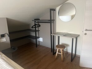 Una habitación pequeña con suelo de madera, un taburete, un escritorio con espejo y una puerta.