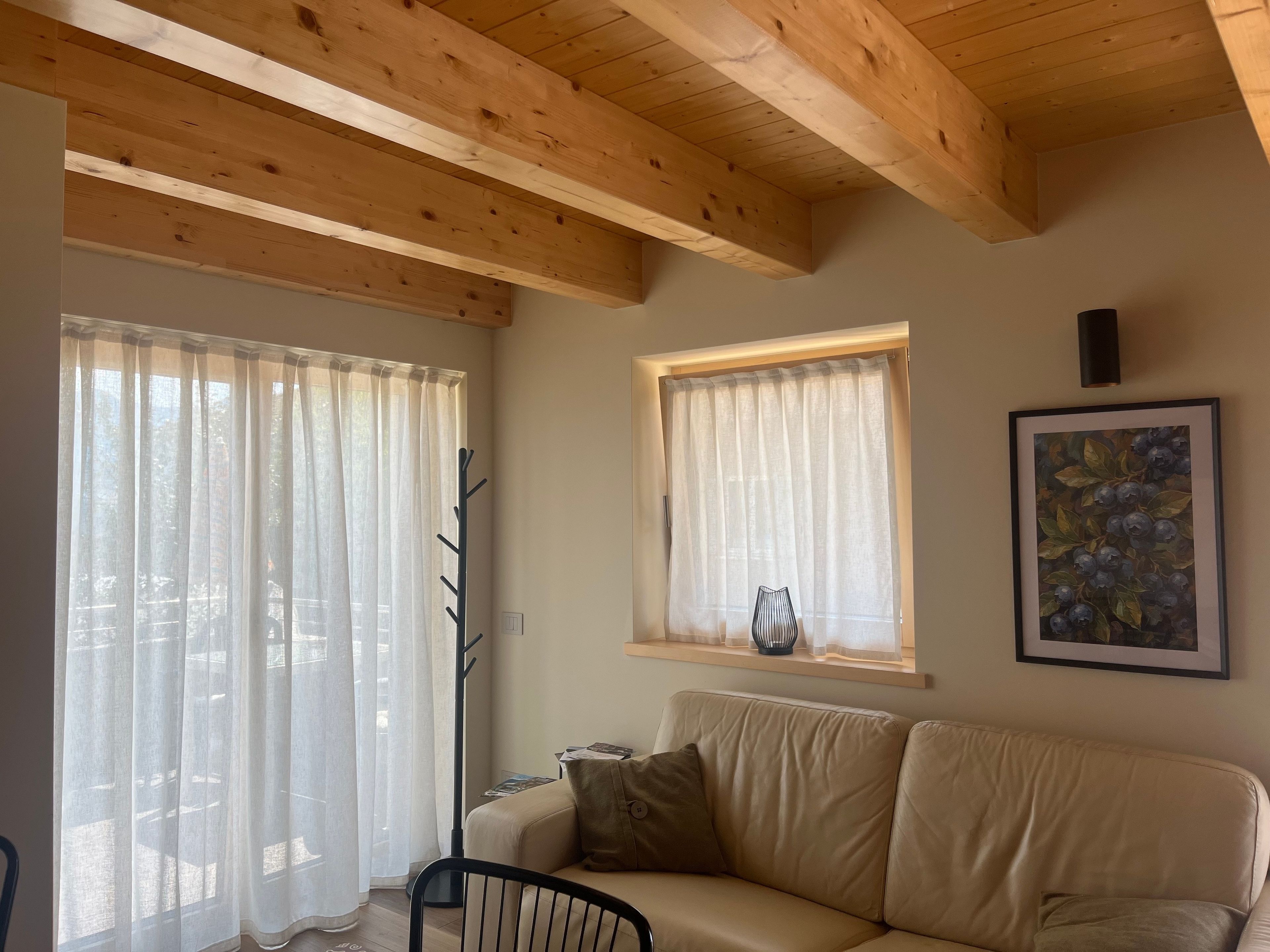 A room with a wooden ceiling, a beige sofa, a window with a vase, and a painting on the wall.