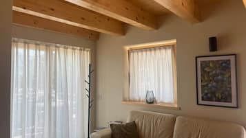 A room with a wooden ceiling, a beige sofa, a window with a vase, and a painting on the wall.
