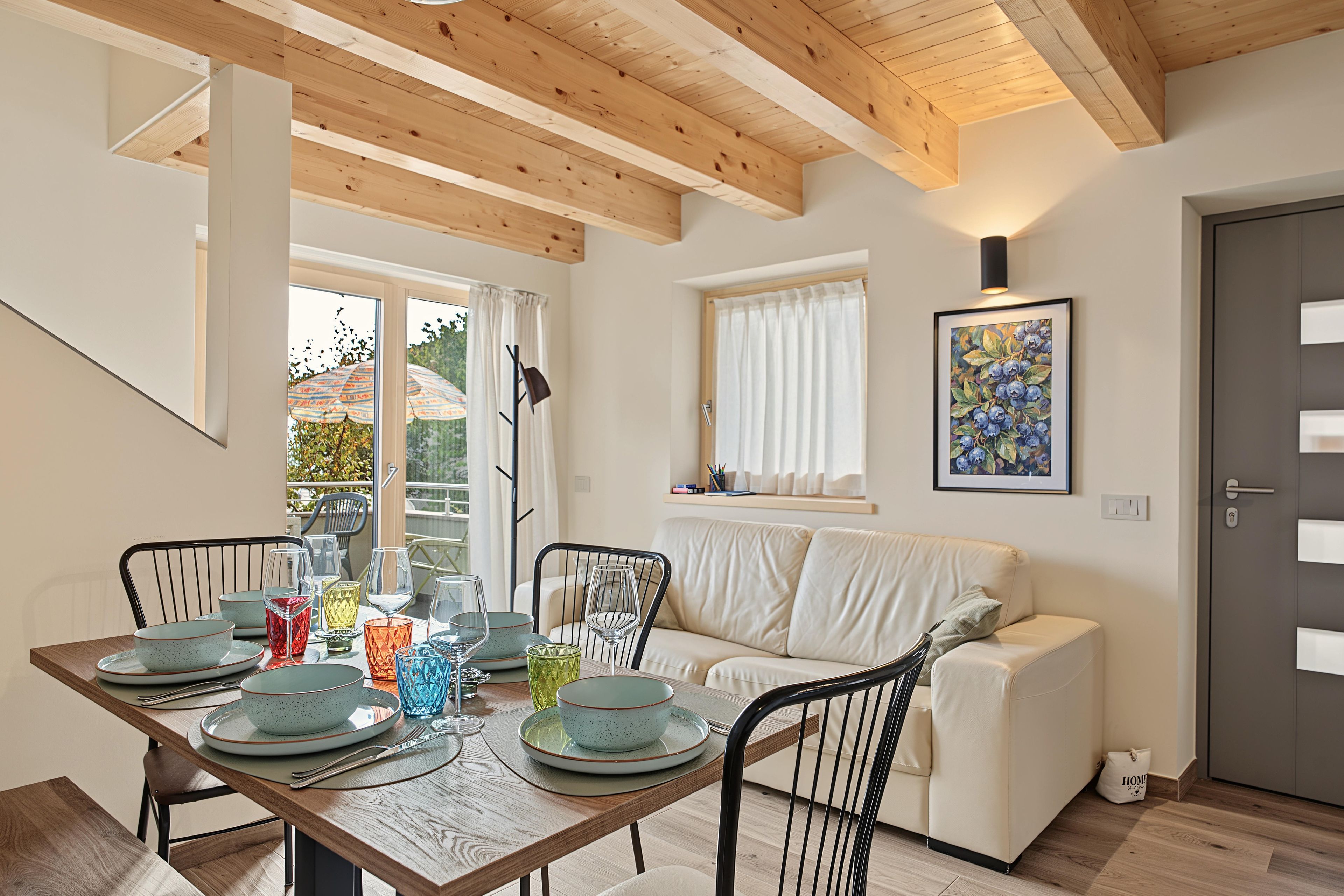 Un comedor con una mesa de madera dispuesta para cuatro, rodeada por sillas, un sofá y una ventana con cortinas.