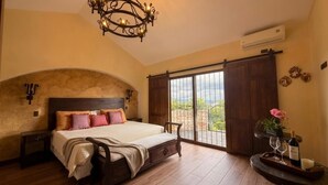Quarto com cama grande, lustre, porta de correr para uma varanda e uma garrafa de vinho sobre uma bandeja.