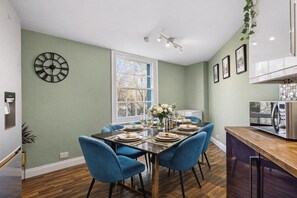 A dining area with a table set for four, blue chairs, a clock on the wall, and a kitchen area with a microwave. - The Penthouse on Bristols Iconic Harbourside (Bristol)
