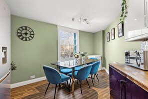 Un coin repas avec une table en verre, des chaises bleues, une horloge au mur et une fenêtre donnant sur un beau paysage.