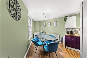 Ein Essbereich mit Tisch und Stühlen, eine Uhr an der Wand und eine Küche mit Schränken und Geräten.