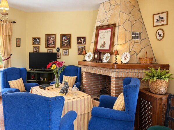 Un salon avec des fauteuils bleus, une cheminée, un téléviseur et divers tableaux encadrés accrochés au mur.