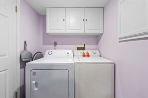 A laundry room with a front-loading washer and dryer, a cabinet above the dryer, and a wall-mounted ironing board. - Castaways 4A 🌊 Gulf Front Condo with Balcony + Pool (Gulf Shores)