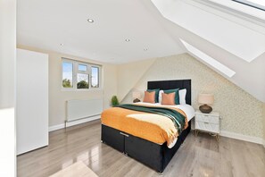 Ein Schlafzimmer mit schrägem Dach, einem Bett mit schwarzem Kopfteil, einem Nachttisch mit Lampe und einem Fenster mit Blick ins Freie.