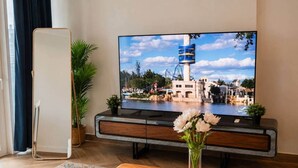 A flat-screen TV on a wooden stand, a potted plant, and a vase with flowers on a wooden table. - 1BR in a Landmark Tower  Rooftop Pool, 360° Views, Near Metro (Dubai)