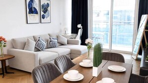 A modern living room with a sofa, dining table, and chairs. There are framed artworks on the wall, a vase with flowers, and a view of the city through large windows. - 1BR in a Landmark Tower  Rooftop Pool, 360° Views, Near Metro (Dubai)
