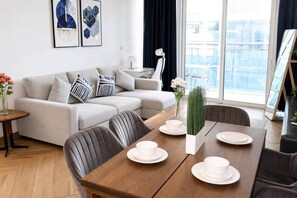 A modern living room with a sofa, dining table, and chairs. There are framed artworks on the wall, a vase with flowers, and a view of the city through large windows. - 1BR in a Landmark Tower  Rooftop Pool, 360° Views, Near Metro (Dubai)