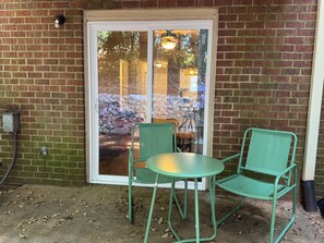 A brick wall with a glass door leading to an outdoor area. There are two green chairs and a small table.