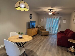 A living room with a ceiling fan, a dining table with chairs, a red sofa, and a television on a wooden cabinet.