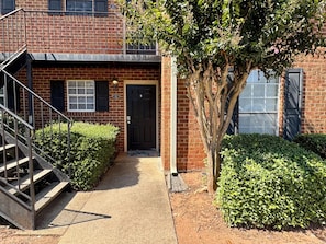 A brick building with a black front door, a small front yard, and a metal staircase leading to an upper level.