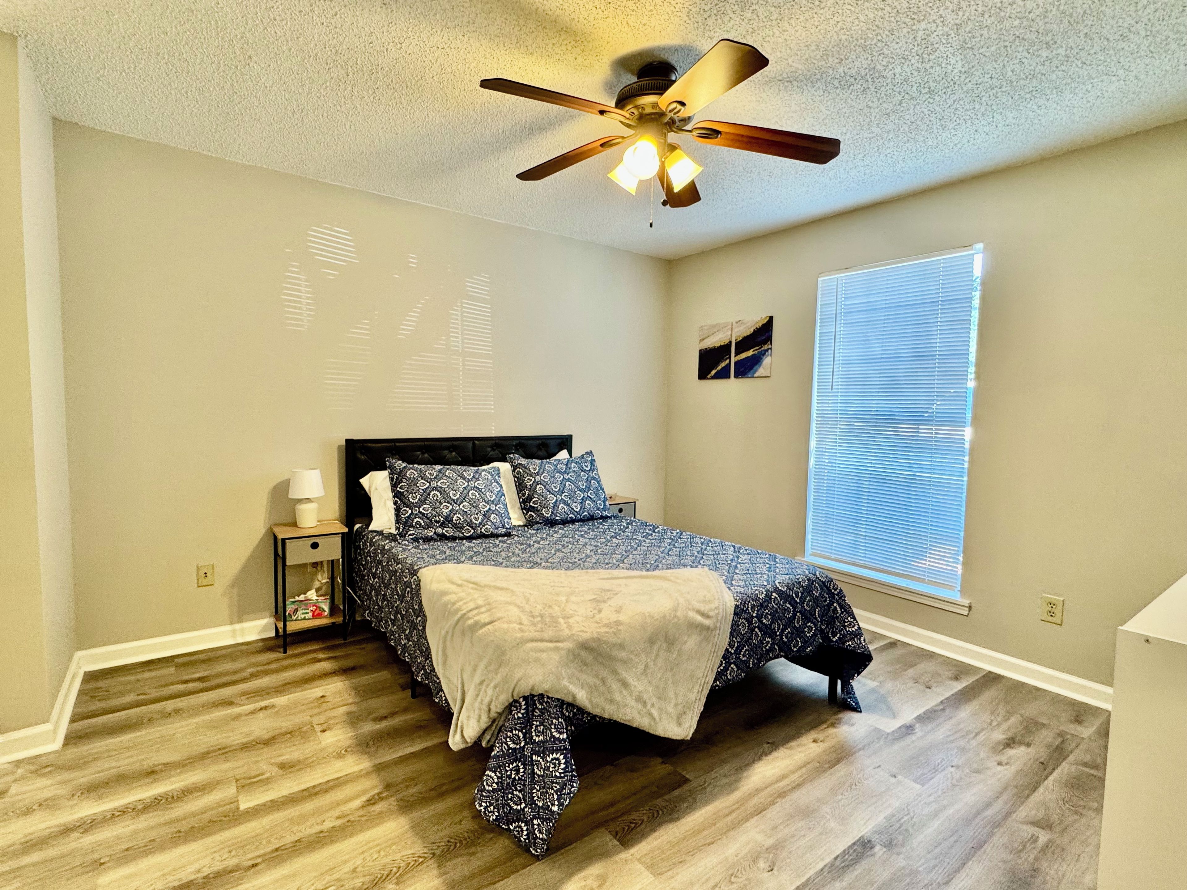 Une chambre à coucher avec un lit, un ventilateur de plafond, une table de chevet, une lampe, une fenêtre avec des stores et un mur orné d’un tableau.