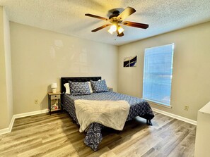 Un dormitorio con cama, ventilador de techo, mesita de noche, lámpara, ventana con persianas y una pared con un cuadro.