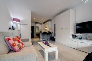 Ein modernes Wohnzimmer mit einer Couch, einem Couchtisch und einem Fernseher. Im Hintergrund ist eine Küchenzeile mit Kühlschrank und Mikrowelle zu sehen.