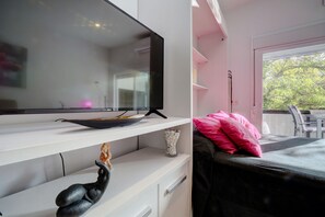 Ein modernes Schlafzimmer mit einem Flachbildfernseher auf einem weißen Regal, einer dekorativen Skulptur, einem Bett mit schwarzem Kopfteil und einem rosafarbenen Kissen.