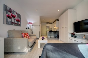 Ein modernes Wohnzimmer mit einer Couch, einem Couchtisch und einem Fernseher. Es gibt eine Küchenzeile mit Kühlschrank und Mikrowelle.