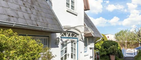 A white house with a sloped roof, a front door with a decorative arch, and a small garden with potted plants.