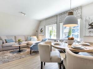 A modern living room with a sofa, dining table, and chairs. There is a window with patterned curtains and a hanging light fixture. - Holiday Apartment Sylt 86 (Sylt)