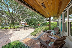 A covered patio area with brick flooring, wooden furniture, and a view of a lush garden with trees and shrubs. - Hibis-Kiss Haven, Heated Pool, Walk To Dune Dog Restaurant (Jupiter)