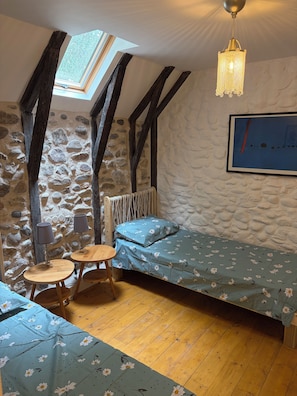 Une pièce chaleureuse avec un plafond en pente, des murs en pierre, un plancher en bois, un lit avec une literie à motifs floraux, une petite table avec des chaises, un luminaire suspendu et un tableau encadré au mur.