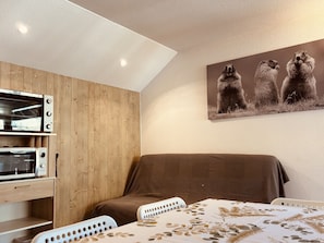 A room with a bed, a table with a leaf-patterned tablecloth, a wall-mounted picture of three animals, and built-in appliances including a microwave and a toaster oven. - Bright studio ski slopes view for (Les Deux Alpes)