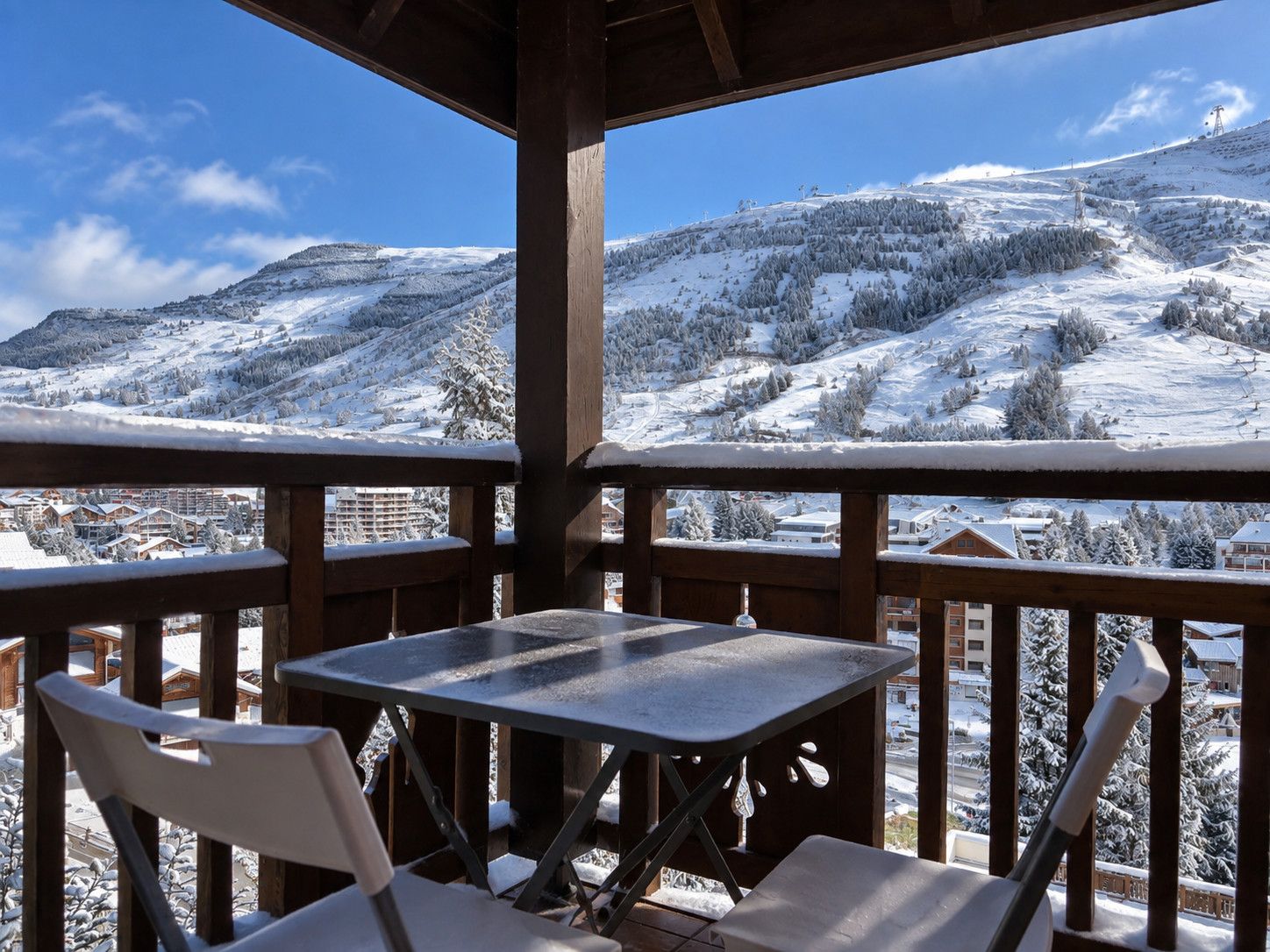Une terrasse en bois avec une table et des chaises, d’où l’on peut admirer un paysage montagneux enneigé, avec des bâtiments visibles au loin.